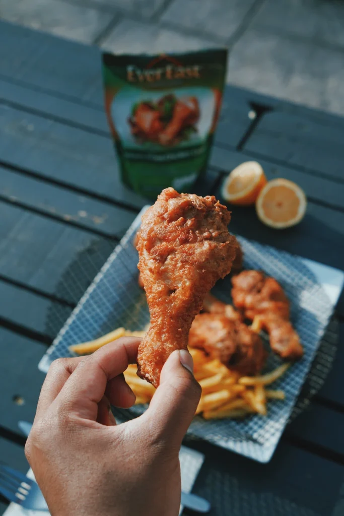 Hand holding a fried chicken drumstick over a plate of fries and chicken on a table