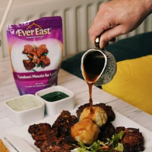 Hand pours sauce from a patterned cup over a plated snack with EverFast Tandoori Masala mix nearby